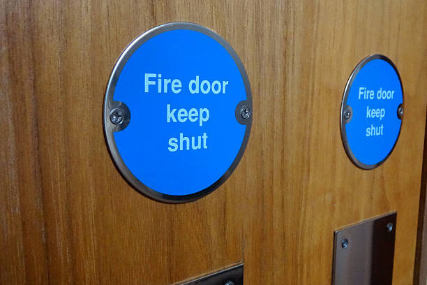 Photo showing a circular blue sign with white writing saying 'Fire Door Keep Shut '.  This safety sign has been screwed onto a wooden fire door.